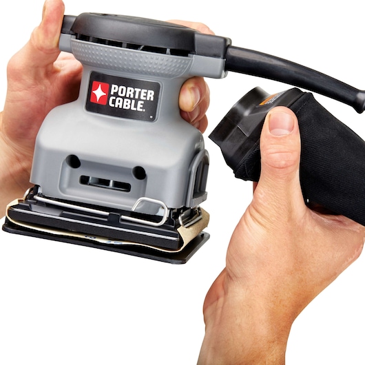 Close-up of hands holding a PORTER-CABLE electric sander, with one hand attaching the dust bag to the tool.