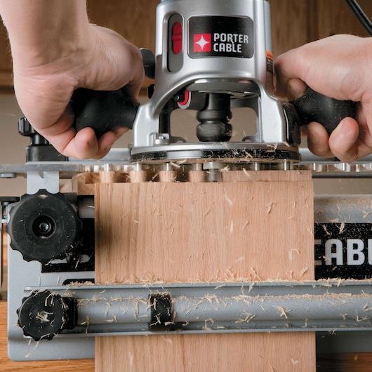 Close-up of a PORTER-CABLE wood router being used to cut a dovetail joint in a piece of wood. The machine is actively shaving wood, and wood shavings are visible around the workpiece. Hands are holding the tool firmly.