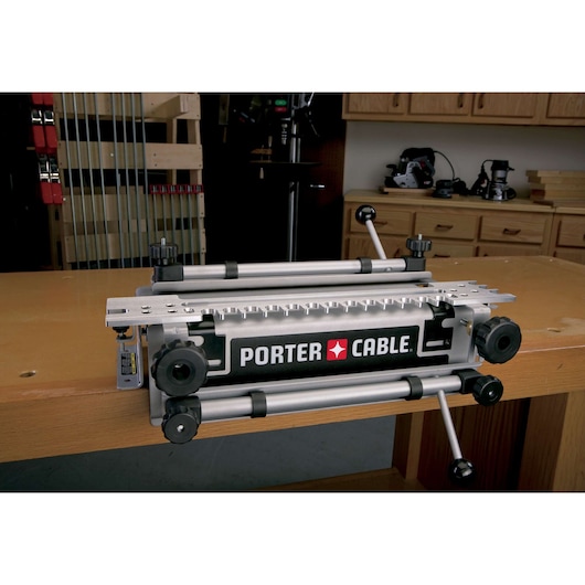 A PORTER-CABLE dovetail jig securely attached to a wooden workbench in a woodworking shop. The jig features multiple adjustment knobs and metal bars for precision joinery work.