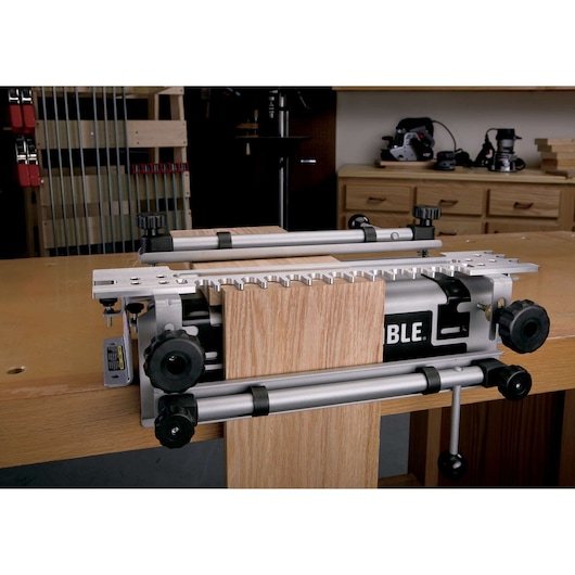 A PORTER-CABLE woodworking jig is attached to a workbench, holding two pieces of wood for precision joinery work. The jig features metal components, adjustment knobs, and a template for cutting dovetail joints. The workshop background includes tools, clamps, and wooden cabinets.