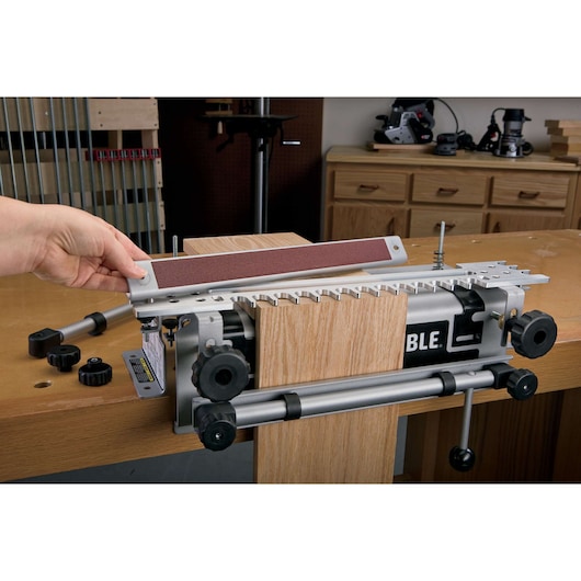 A PORTER-CABLE model 4210 woodworking jig is clamped to a workbench, holding two pieces of wood. A hand is adjusting the removable guide on the jig. Several tools and a wood shop background are visible.