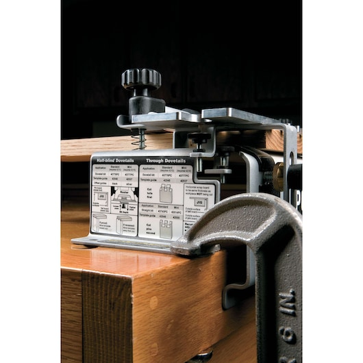 A PORTER-CABLE 4212 dovetail jig clamped to a wooden workbench. The metal jig includes various adjustment knobs and a detailed instruction chart for half-blind and through dovetail joinery visible on the front. A large metal clamp secures the jig and wood in place for woodworking.
