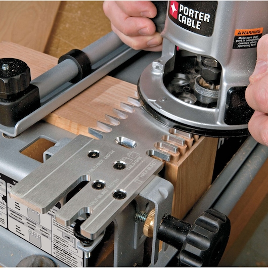 Close-up of a PORTER-CABLE woodworking tool being used to cut dovetail joints in a piece of wood, with hands operating the device and metal jig guiding the cuts.