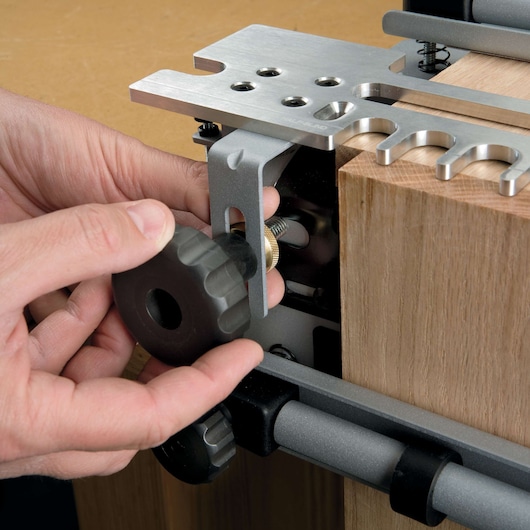 Close-up view of a person's hands adjusting the knob on a PORTER-CABLE dovetail jig attached to a wooden board. The metal components and adjustment mechanism are visible.
