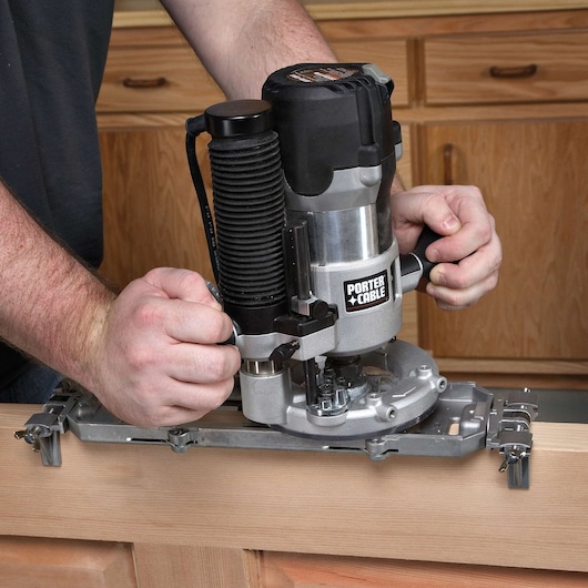 A person uses a PORTER-CABLE router tool to work on a piece of wood in a workshop with wooden cabinets in the background.
