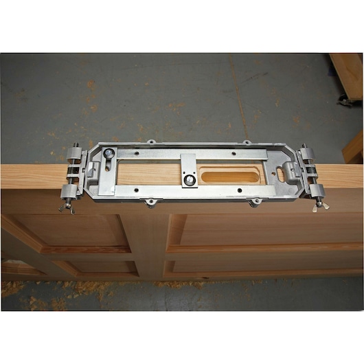 A metal door hinge mortise jig is clamped onto the edge of a wooden door, allowing for precise routing of hinge slots. Wood shavings are visible on the ground below the door.