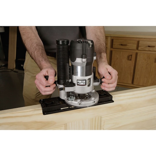 A person using a PORTER-CABLE plunge router to cut or shape a piece of wood on a workbench, with cabinets visible in the background.