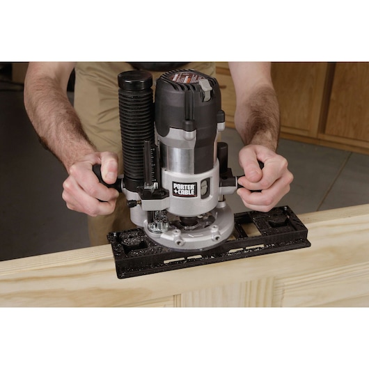 A person is using a PORTER-CABLE plunge router to cut or shape wood. The tool is being guided along the edge of a light wooden board, and wood shavings are visible around the base of the router.