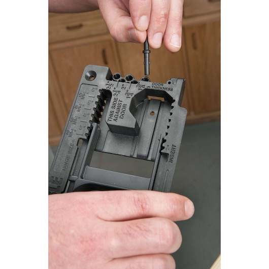 Close-up of a person adjusting a PORTER-CABLE door hardware template, used for marking and drilling precise holes for door locks and hinges. The person is holding a black plastic template and inserting a pin to set the door thickness. Cabinet doors and a work surface are visible in the background.
