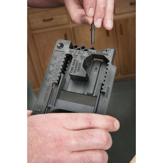 A close-up of a person using a PORTER-CABLE photography tool, placing a metal pin into a black plastic guide, with labeled slots for door thickness and hinge size. Wooden cabinets are visible in the background.