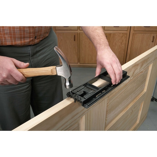 A person uses a hammer and a black door hardware jig to install or mark a location on a wooden door, with cabinet doors visible in the background.