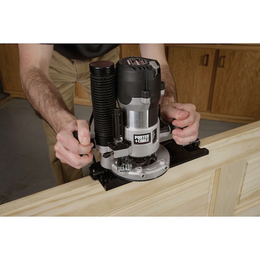 A person is using a PORTER-CABLE wood router to work on a wooden surface, with hands gripping the tool for precision. The router is positioned on the edge of the wood, likely for shaping or trimming.
