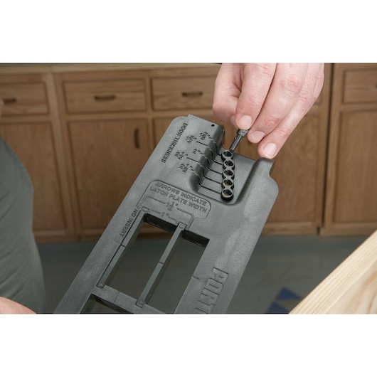 A close-up of a hand adjusting a PORTER-CABLE door lock installation kit, showing detail of its settings and measurements for door hardware installation. The black plastic tool is held above a work surface in a workshop with wood cabinets in the background.