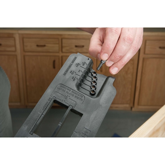 A close-up of a person adjusting a PORTER-CABLE door lock installation kit in a woodworking setting. The person is holding a small tool above the black plastic template, which has labeled holes for door thickness measurements. Wooden cabinets are visible in the background.