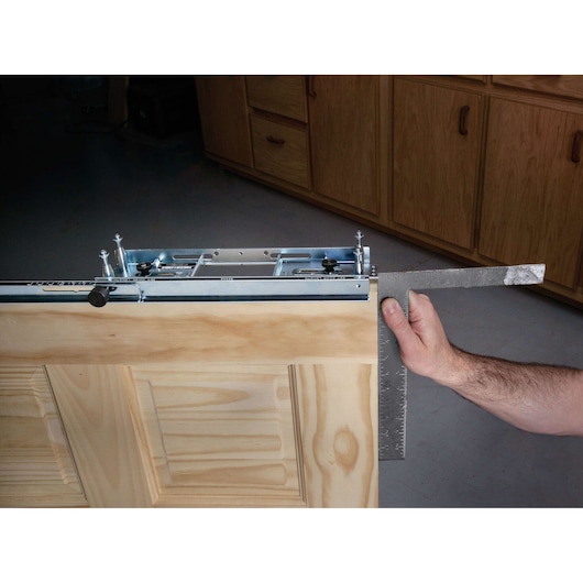 A person is holding a metal square ruler against the edge of a wooden door panel, with a PORTER-CABLE door hinge template jig attached on top of the door. Wooden cabinets are visible in the background.