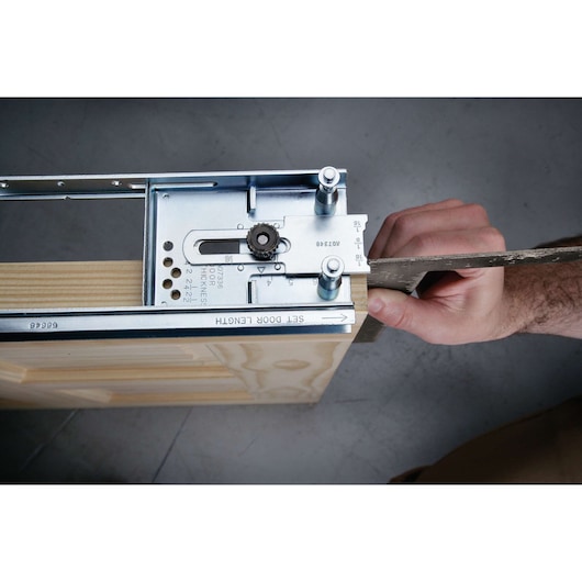 Close-up of a person adjusting a metal hardware jig tool attached to the top of a wooden door, likely used for setting door length or installing door hardware.