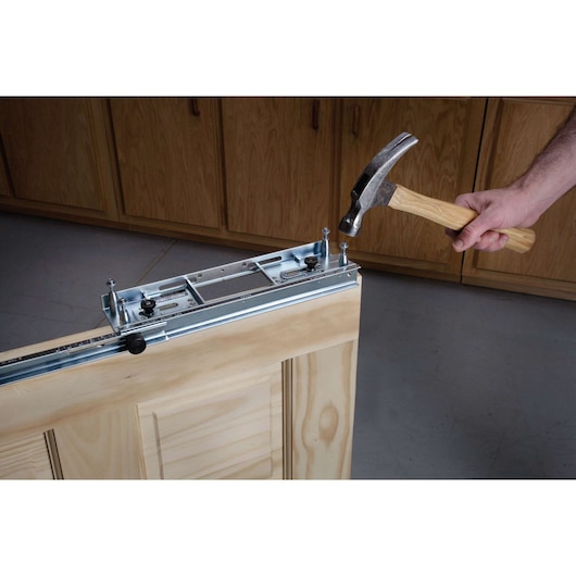 A person is using a hammer to install a metal hardware track onto the top edge of a wooden door, with wooden cabinets visible in the background.
