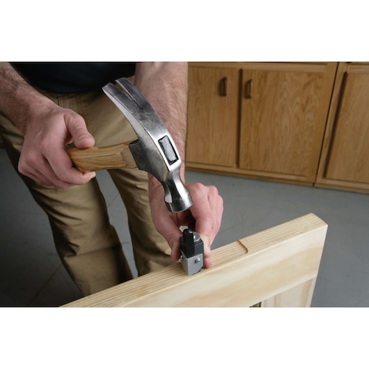 A person using a hammer to strike a metal tool positioned on the edge of a wooden board, likely for woodworking purposes. Wooden cabinets are visible in the background.
