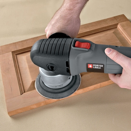 A person using a PORTER-CABLE electric sander to smooth the surface of a wooden cabinet door.
