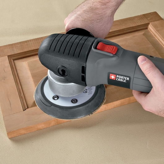 A person using a PORTER-CABLE power tool to sand or polish a wooden frame on a beige surface.