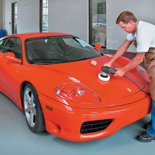 A person using a PORTER-CABLE 7346SP polisher on the hood of a bright red sports car inside a garage or workshop. The person's face is blurred for privacy.