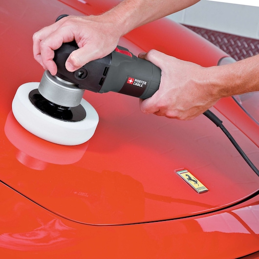 A person is using a PORTER-CABLE power tool with a sponge pad to polish the surface of a red car. The car features a visible emblem on its hood.