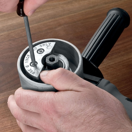 Close-up of a person using a screwdriver to adjust or secure a metal plate on a power tool, likely an angle grinder, with a black handle and part of a gray housing visible.