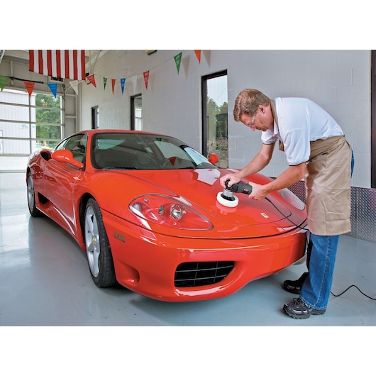 A person using a PORTER-CABLE 7424XP polisher on the hood of a shiny red sports car inside a garage. The person’s face is blurred and they are wearing an apron and jeans.
