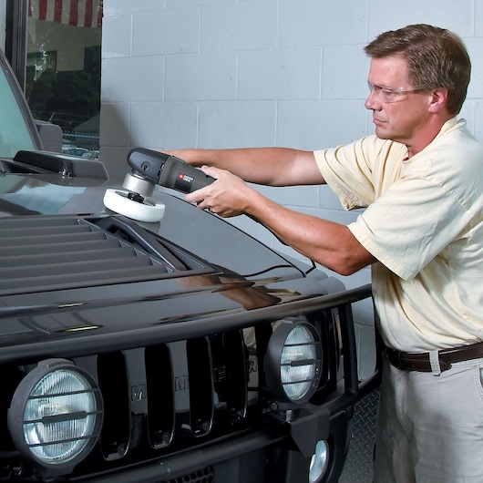 A person uses a PORTER-CABLE 7424XP polisher to polish the hood of a black vehicle inside a garage. The person's face is blurred.
