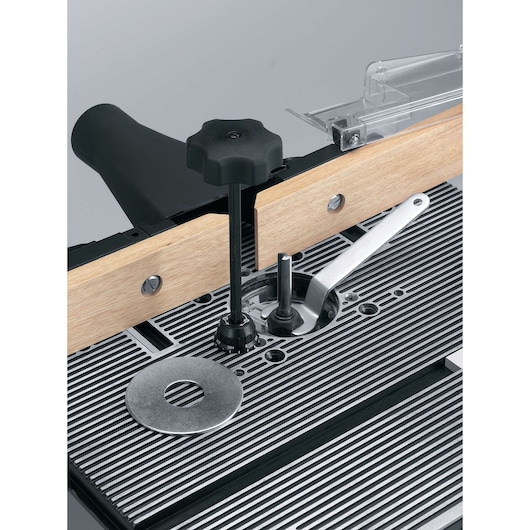 Close-up view of a router table with a metal fence, adjustment knob, and dust port. The tabletop has grooves and a round metal insert for woodworking.