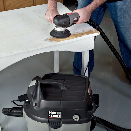 A person is using a PORTER-CABLE power tool to sand a white wooden table, with a PORTER-CABLE shop vacuum visible nearby to collect dust.