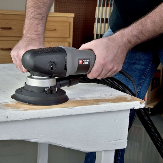 A person using a PORTER-CABLE random orbital sander on a white wooden table, sanding the surface. The sander is connected to a vacuum hose. The scene takes place in a workshop setting.