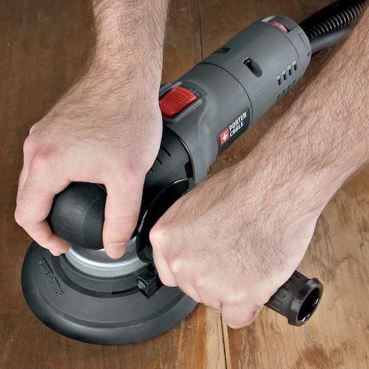 A PORTER-CABLE electric sander being held and operated by two hands on a wooden surface.