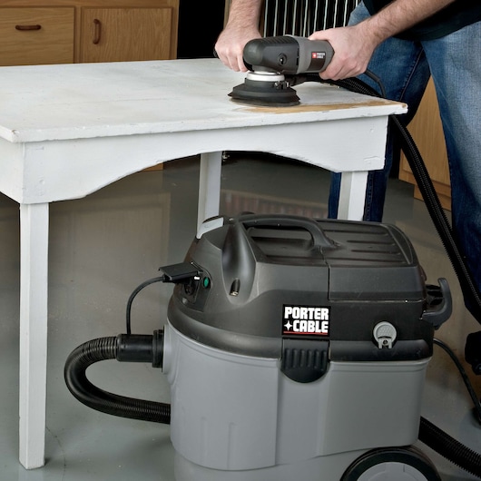 A PORTER-CABLE branded wet/dry vacuum is placed on the floor next to a person sanding a white wooden table using a power sander. The vacuum’s hose is connected and the scene takes place indoors, possibly in a workshop setting.