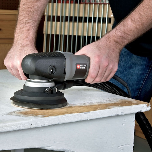 A person is using a PORTER-CABLE electric sander to refinish a wooden surface, attached to a dust collection hose. The hands are visible holding the tool over the edge of a table.
