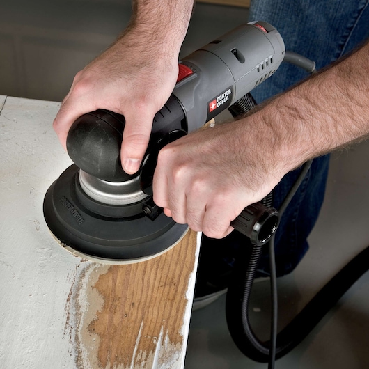 A person is using a PORTER-CABLE electric sander to smooth a wooden surface, removing old paint from a piece of wood furniture.