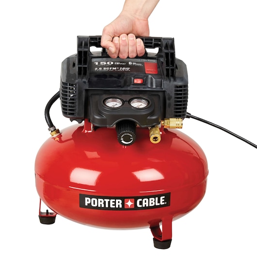 A PORTER-CABLE C2002 red and black portable air compressor being held by a hand on the handle. The compressor has pressure gauges, a power switch, and connection ports visible on the front.