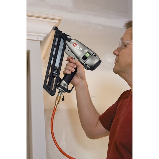 A person using a PORTER-CABLE DA250C pneumatic nail gun to attach trim to a wall, with the tool connected to an air hose. The person's face is obscured.