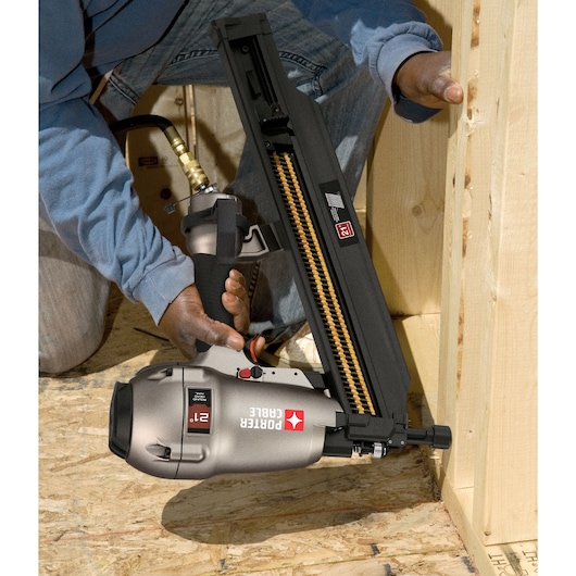A person using a PORTER-CABLE FR350B pneumatic framing nailer to fasten wooden boards during construction. The nailer, connected to an air hose, is positioned against the wood frame.