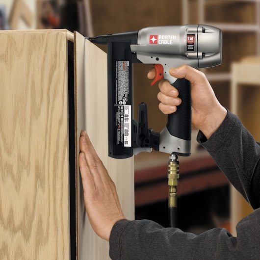 A person uses a PORTER-CABLE NS150C pneumatic nailer to attach a panel to a wooden cabinet in a workshop.