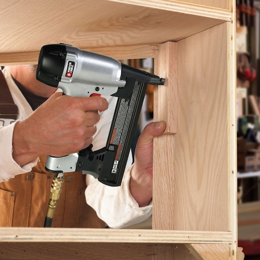 A person is using a PORTER-CABLE pneumatic nail gun to fasten wooden boards together in a woodworking project. The nail gun is being held close to the wood surface, and the person is wearing a brown work apron.