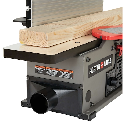 Close-up of a PORTER-CABLE jointer, showing a piece of wood placed on the machine's flat surface. The jointer has safety warning labels, a dust collection port, and the brand logo clearly visible.