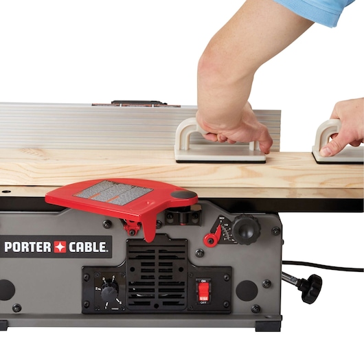 Close-up of a PORTER-CABLE jointer (SKU: PC160JT) in use, with two hands holding push blocks to guide a wooden board through the machine.