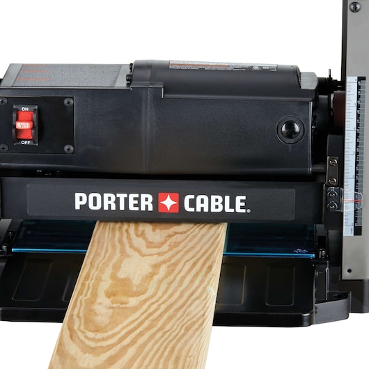 A PORTER-CABLE wood planer with a piece of wood passing through the machine. The planer has a black exterior and visible control switches on the left side.