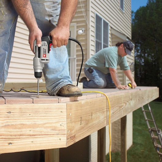 A person using a PORTER-CABLE electric drill to install screws into a wooden deck, with another person in the background measuring or working on the deck. The deck is attached to a house and outdoor construction is underway.