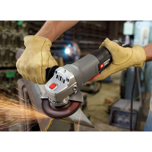 A close-up view of a person wearing yellow work gloves using a PORTER-CABLE angle grinder to grind a piece of metal, with sparks flying. The scene takes place in a workshop with industrial equipment in the background.