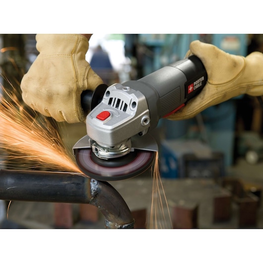 A person wearing protective gloves uses a PORTER-CABLE angle grinder to grind a metal pipe, producing sparks in a workshop.
