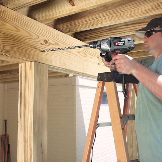 A person using a PORTER-CABLE PC700D electric drill with a long auger bit to drill through a wooden beam, standing on a ladder in a construction or woodworking setting. The person’s face is obscured.