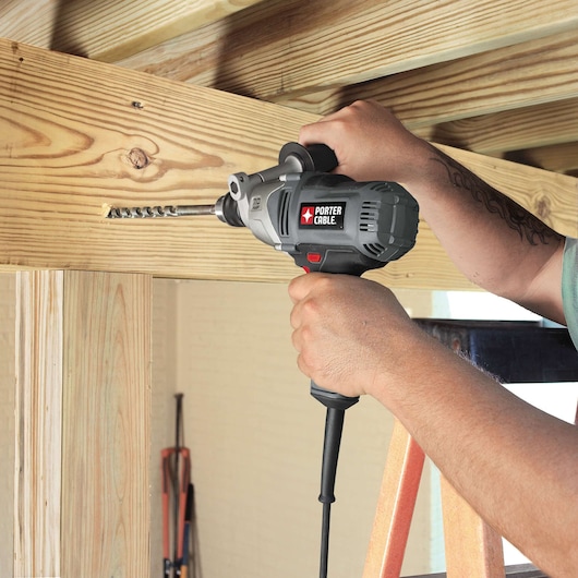 A person using a PORTER-CABLE corded drill (model PC700D) to drill a hole in a wooden beam during construction work.