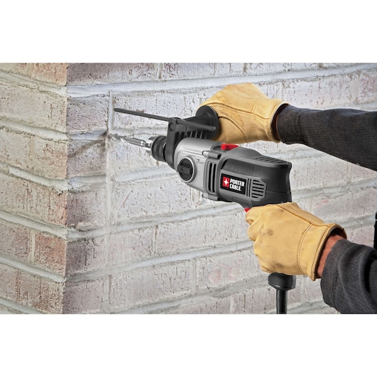 A person wearing yellow work gloves is using a PORTER-CABLE hammer drill to drill into a brick wall. Dust is visible as the drill bit penetrates the mortar between the bricks.